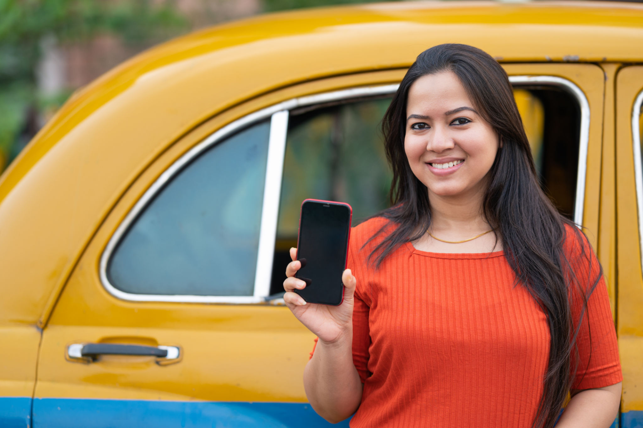 Outdoor image of young Woman showing mobile phone while standing near yellow taxi. Online taxi booking, Taxi booking app on smartphone.